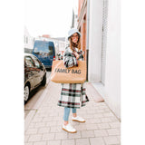 Woman holding a large, textured beige "FAMILY BAG" tote with black text, wearing a plaid coat.