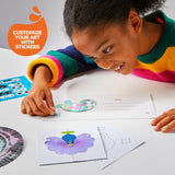 Smiling child customizing a white spin art card with a colorful sticker, surrounded by art supplies.