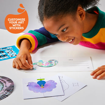 Smiling child customizing a white spin art card with a colorful sticker, surrounded by art supplies.