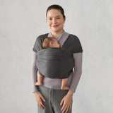 Smiling woman wearing a grey Ergobaby Aura knit wrap with a sleeping baby nestled inside.