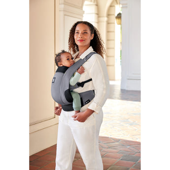 Woman carrying baby in a grey Ergobaby Away carrier with black accents, walking outdoors.