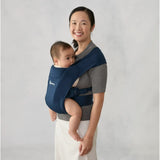 Smiling woman wearing a navy Ergobaby Embrace newborn carrier with a baby facing inward.