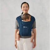 Woman wearing a navy Ergobaby Embrace carrier with a baby sleeping against her chest.