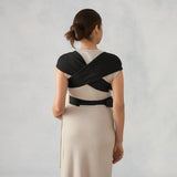 Woman from behind wearing a black Ergobaby Embrace carrier over a light dress, showing the crisscross back straps.