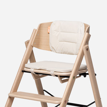 KAOS Klapp reversible highchair cushion set in cream, shown on a natural wood highchair.