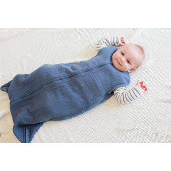 Happy baby in a blue Lulujo 100% cotton muslin sleep sack on a white textured blanket.
