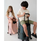 Two children riding on 24-inch MiaMily check-in suitcases: a girl on a pink one, a boy on a dark green one.