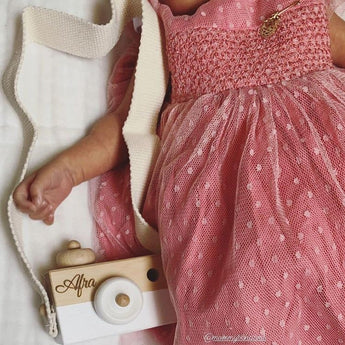 Personalized wooden camera toy with "Afra" engraved, white lens, and cream strap, beside a baby in a pink polka dot dress.
