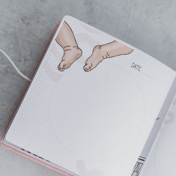 Open pink suede baby milestone album page with "DATE" field, baby feet illustration, and photo circle.