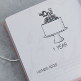 Open pink suede baby milestone album page with "is One" cake illustration and "1 YEAR" text.