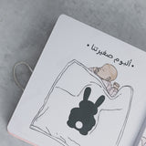 White baby milestone photo album with sleeping baby and black bunny illustration, Arabic text.