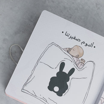 White baby milestone photo album with sleeping baby and black bunny illustration, Arabic text.