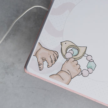 Pink suede baby milestones photo album with a cute illustration of baby hands holding a wooden bird rattle.