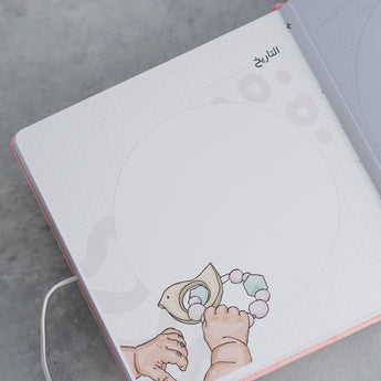 Open pink suede baby milestone album page with "التاريخ" (Date) and baby hands holding a pastel rattle illustration.