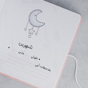 Open pink baby milestone album page with a moon and stars illustration and Arabic text.