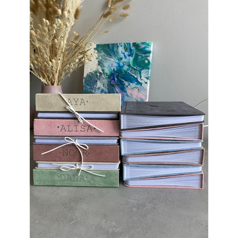 Stack of personalized suede baby milestone photo albums in beige, pink, brown, and green, with a dark gray album open.