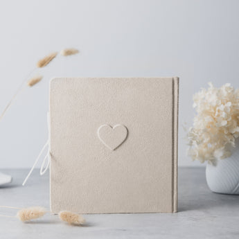 Beige suede baby milestones photo album with an embossed heart, tied with a white ribbon.