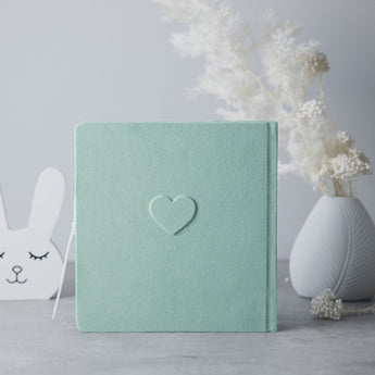 Mint green suede baby photo album with an embossed heart, next to a white bunny decor and vase.