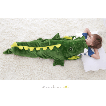 Green crocodile-shaped kids sleeping sack with yellow accents, a child sleeping inside on a white fluffy rug.