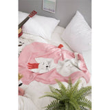 Cozy pink knit blanket with a white cat design, red detail, and tassel, on a bed with pillows and a guitar.