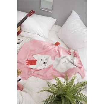Cozy pink knit blanket with a white cat design, red detail, and tassel, on a bed with pillows and a guitar.