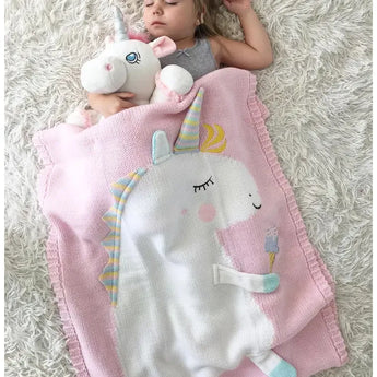 Sleeping child with a pink knit unicorn blanket and a white unicorn plush toy on a fluffy white rug.