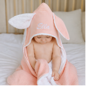 Personalized organic peach bunny hooded towel with white trim, embroidered "Ella" on the hood, worn by a baby.