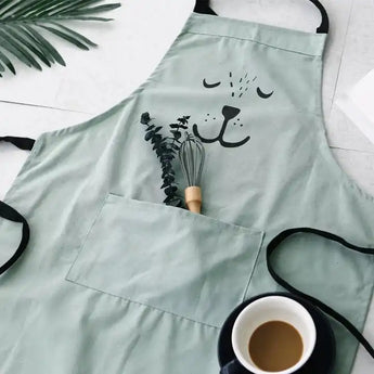 Light green kids bear apron with black straps, front pocket holding a whisk and plant, next to a coffee cup.