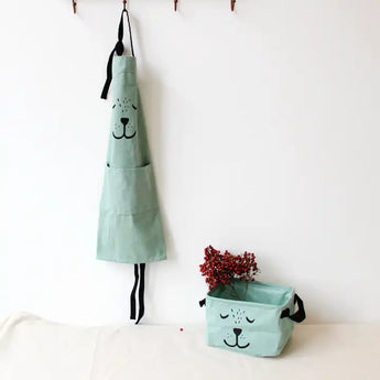 Light green kids bear apron with black ties, hanging next to a matching storage basket with red berries.