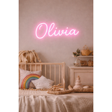 Pink neon LED sign "Olivia" above a wooden crib with rainbow pillow, beige bedding, and decor.