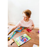Young boy with red hair creating OOLY Foil-tastic art on a wooden table, featuring colorful foil sheets and whimsical designs.