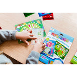 Child's hands applying silver foil to a dragon art card from the Ooly Foil-Tastic set on a wooden table.