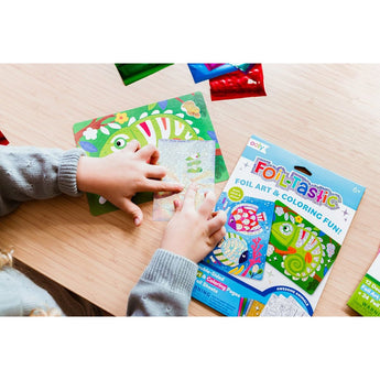 Child's hands applying silver foil to a dragon art card from the Ooly Foil-Tastic set on a wooden table.