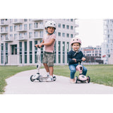 Two children with helmets, one standing on a white Scoot & Ride Highwaykick 1 scooter, another sitting on a pink one.