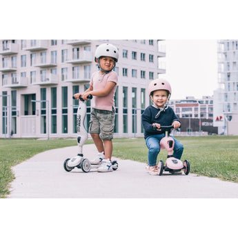 Two children with helmets, one standing on a white Scoot & Ride Highwaykick 1 scooter, another sitting on a pink one.