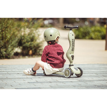Child in a green helmet sitting next to a sage green Scoot & Ride Highwaykick 1 2-in-1 scooter.