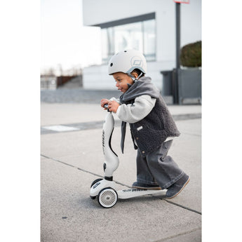 Toddler in a white helmet and gray vest riding a white Scoot & Ride Highwaykick 1 scooter outdoors.