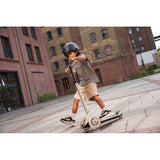 Kid in helmet riding a beige Scoot & Ride Highwaykick 3S LED 3-wheel light-up scooter outdoors.