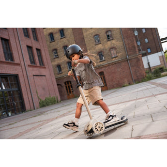 Kid in helmet riding a beige Scoot & Ride Highwaykick 3S LED 3-wheel light-up scooter outdoors.