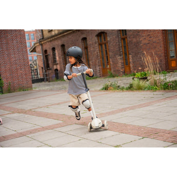 Child in helmet and pads riding a white Scoot & Ride Highwaykick 3S LED 3-wheel scooter outdoors.