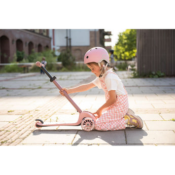Girl in pink helmet and checkered overalls adjusting a pink Scoot & Ride Highwaykick 3S LED 3-wheel scooter.