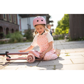 Young girl in pink helmet and checkered overalls sitting next to a pink 3-wheel light-up scooter.