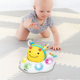 Baby crawling towards a yellow and blue Skip Hop Follow Bee toy with colorful lights on a grey playmat.