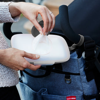 Hands pulling a white wipe from a translucent Skip Hop Grab & Go Wipes Case with a dark gray lid, next to a stroller.