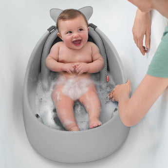 Happy baby in a gray Skip Hop Moby 3-stage tub with mesh sling and bubbles, being bathed by an adult.