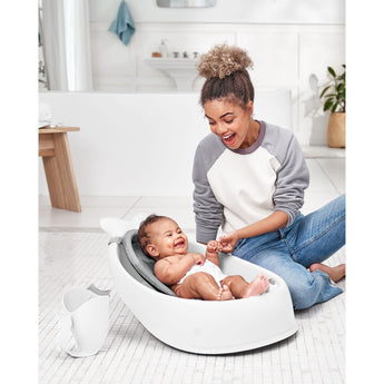 Happy mom bathing baby in a white Skip Hop Moby bath tub with a gray mesh sling and white rinser.
