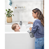 Smiling woman rinses baby's hair with a white Skip Hop Moby Waterfall Rinser in a white bathtub.