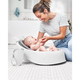 Smiling mom interacting with baby in a white Skip Hop Moby bath rinser, with a grey mesh seat.
