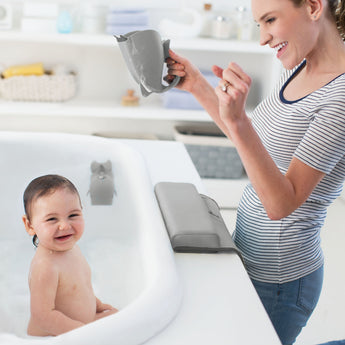 Smiling baby in a white tub, happy mom holding a gray Skip Hop Moby Waterfall Rinser with a soft lip.