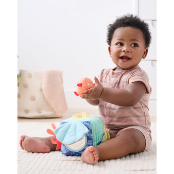 Happy baby playing with a soft, multi-textured Skip Hop Seascape Activity Cube and an orange puffer fish rattle.